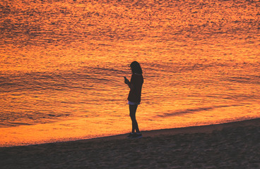 Junges Mädchen, junge Frau alleine am Strand mit ihrem Smartphone im goldenen Licht direkt nach dem Sonnenuntergang – low Key © redaktion93