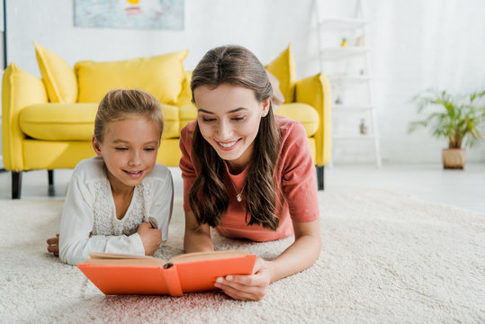happy babysitter lying on carpet with cheerful kid while reading book