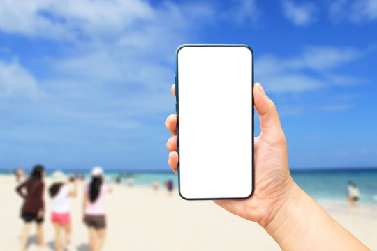 Mock up image of A hand holding a blank screen of smartphone on the beach and sea blurred​ background.