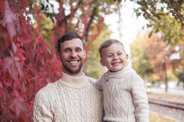 Dad and mvlenky son walk in the autumn park. Bright, warm autumn. Red leaves. Cozy. Portrait of dad and son in knitted sweaters.