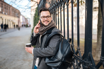 Fototapeta premium Portrait of elegant young man with backpack and takeaway coffee talking on mobile phone.