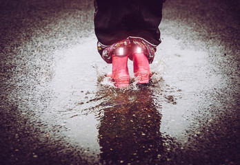 Girl having fun, jumping in water puddle on wet street, wearing rain boots with reflective detail...