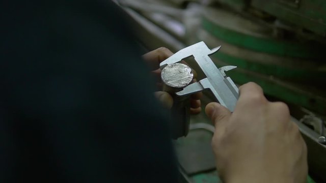 The hands of a factory worker are holding a metal detail and slide gauge. The worker is using the hand caliper to measure the detail. Then he turns the detail upside down and measures it again.
