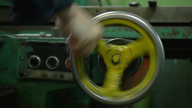 A factory worker is spinning with his fist a metal wheel of some appliance on a plant. We can see a lot of gear and heavy machinery around.