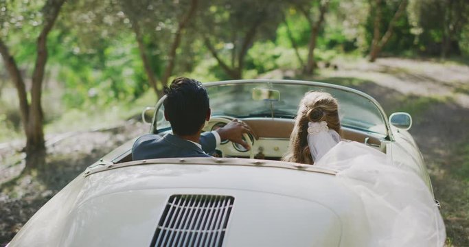 Happy diverse newly wed couple eloping on a sunny country road, bride and groom driving together in a white vintage convertible