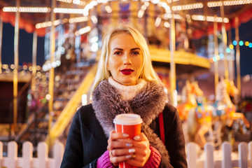 Picture of young woman with glass of coffee in hands on walk on background of carousel