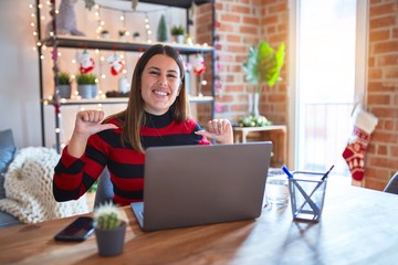 Beautiful woman sitting at the table working with laptop at home around christmas lights looking confident with smile on face, pointing oneself with fingers proud and happy.