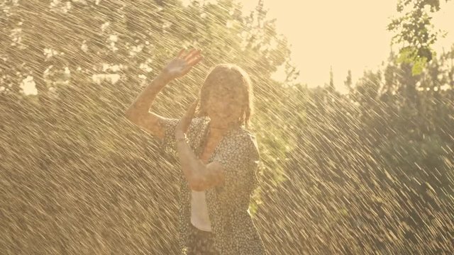 Excited young brunette woman jumping near the sprinkler and becoming wet while having fun in the park