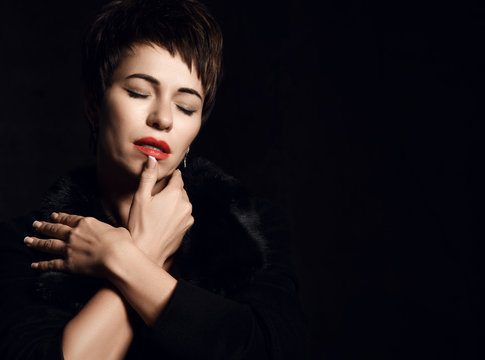 Portrait of sensual woman with short haircut posing with closed eyes touching her lips with her thumb on dark