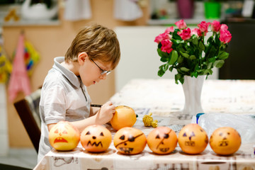 Little kid boy making Jack-o-lantern with drawing scary faces on mandarine, tangerine or clementine. Happy child making preparation for Halloween party at home, indoors