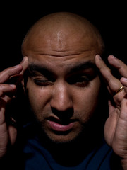 Low key photo of a brown bald headed man placing both hands on forehead due to a headache