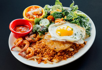 Close up image of Asian pineapple fried rice with fried egg, shrimp, and vegetables salad on a white plate and black background