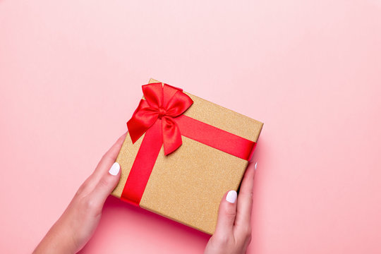 Woman manicured hands holding red and golden wrapped present or giftbox on pastel pink background, copy space, top view, flat lay. Background for Valentine's Day, Mother's Day.