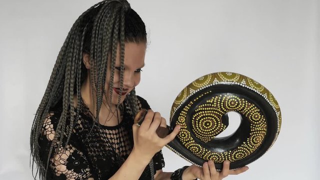 A gothic girl with long grey afro braids and a dark make up is looking at a didjeridu and laughing. A creative concept for a music gear store or a music intsruments shop.