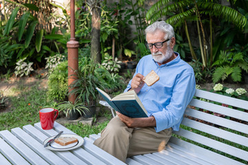 Senior elderly man eating breakfast with mug of coffee reading book in garden