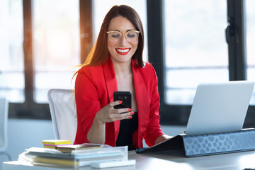 Smiling young businesswoman using her mobile phone while working with computer in the office.