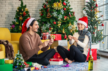 Obraz na płótnie Canvas Happy couple in santa hat sitting on the floor and holding lighting sparkler sticks. Young beautiful woman and man enjoy and celebrate family Christmas party with decoration Christmas tree background.