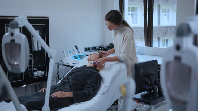 Young woman receiving non-surgical rejuvenation facial treatment lying on couch. Cosmetologist in beauty spa salon performing ultrasonic face skin massage on customer. Rf-lifting.