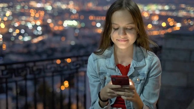 Smiling cute teenage girl communicates on a mobile phone in social networks. Israeli teen femle with night Haifa in background