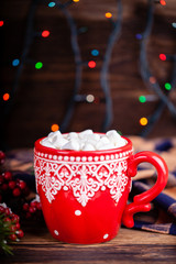 Red mug with ornament with hot cocoa drink and sweet marshmallows in cozy decorations with Christmas garland lights background. New Year holidays concept.