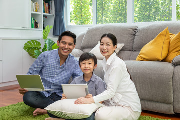 Happy Asian parent sitting together with boy on floor in living room; father working on laptop, and son using tablet with mother; looking at camera
