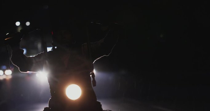 Biker riding his chopper motorcycle with high handlebars on a highway at night in the rain. Viewed from his front in close-up, silhouetted by bright headlights of a car behind him in background