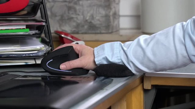 Ein Arbeitsplatz mit Computer. Auf dem Schreibtisch arbeitet eine Hand in entlastender Position mit einer ergonomischen Maus. Neben der vertikalen Maus unterst&uuml;tzt ein kleines Kissen das Handgelenk. 