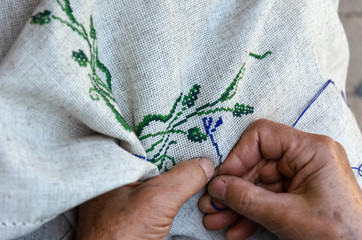 Hands of an elderly woman embroidering a cross-stitch floral pattern on linen fabric. Embroidery, handwork, needlecraft concept. Closeup - obrazy, fototapety, plakaty