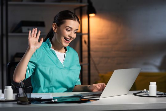 Attractive Nurse In Uniform Sitting At Table And Having Video Chat During Night Shift