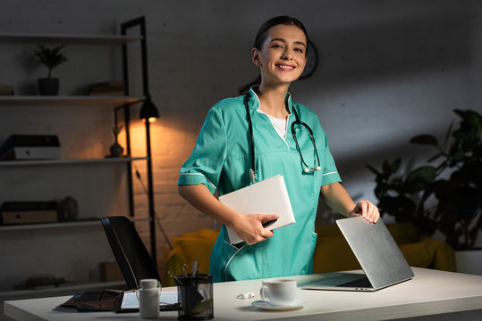 Smiling Nurse In Uniform Holding Notebook And Closing Laptop During Night Shift