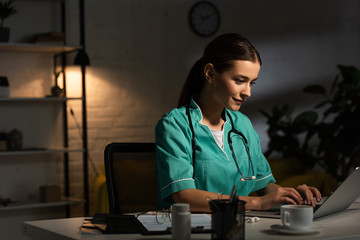 attractive nurse in uniform sitting at table and using laptop during night shift