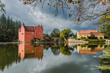 Cervena Lhota, Czech Republic - September 28 2019: View of famous red castle standing on a rock in the middle of a lake. Sunny day and blue sky with clouds. Reflection of buildings in water.