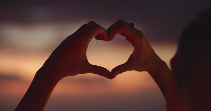 Inspired young woman holding up heart shape at majestic sunset close-up. Back of blonde girl showing symbol of love gesture admiring sumer scenery.