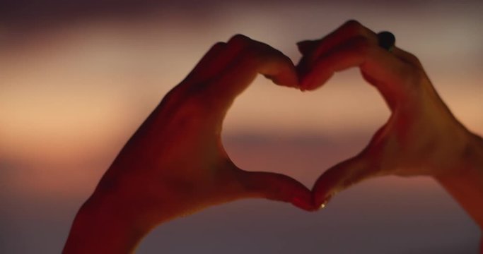 Close-up hands of woman aking heart shape love symbol against beautiful colorful sunset sky. Concept of love and freedom.