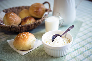Buns in a wicker basket, cottage cheese in a cup and milk on a table covered with a tablecloth.