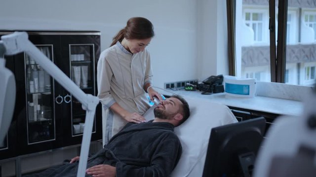 Positive female beautician doing refreshin skincare facial massage on man's face. Relaxed bearded man lying on the couch enjoying ultrasound healthy rf-lifting procedure at salon.