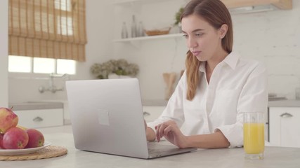 Confident Caucasan businesswoman typing on the laptop at home and drinking orange juice. Young pretty girl in white shirt working online from home. - Powered by Adobe