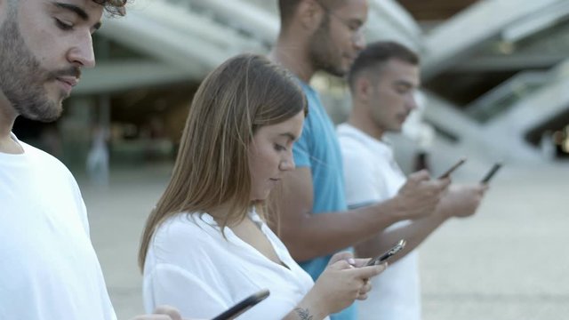 Thoughtful people standing in row and using digital devices. Focused young friends using smartphones on street. Technology concept