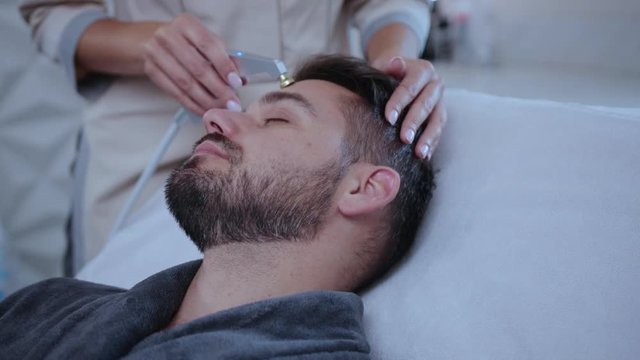 Young man relaxing in beauty salon receiving ultrasonic lubrication face skin care lifting procedure lying on coach. Hands of professional beautician using hardware cosmetology.