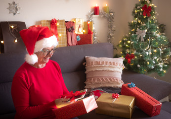 A pleasant surprise. The old lady in a Santa Claus hat receives a new tablet as a Christmas present. One senior people sitting on the sofa. Christmas tree and gifts in the background