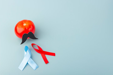 Blue and red ribbons and tomato with black fake paper moustaches on a blue background as a symbol of prevention prostate cancer awareness, copy space. Men's health and a world cancer day