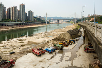 Chongqing, China - March 17, 2018: Trucks, working equipment on the river bank. In the distance is a bridge across the river, city blocks and highways. Jialing River in Chinese city of Chongqing.