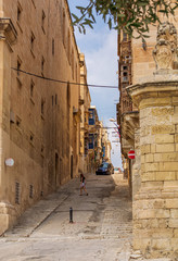 Young girl crossing typical Valletta street with rising road with stairs and traditional Maltese wooden balconies in the distance.