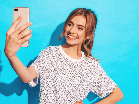 Portrait of cheerful young woman taking photo selfie. Beautiful girl holding smartphone camera. Smiling model posing near blue wall in studio
