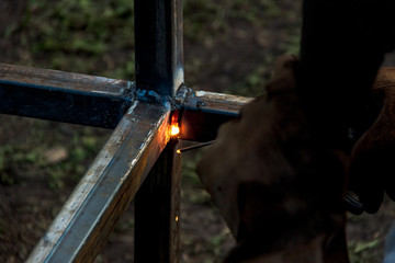 A strong man is a welder, welding mask and welders leathers, a metal product is welded with a arc welding machine at the construction site, blue sparks fly to the sides
