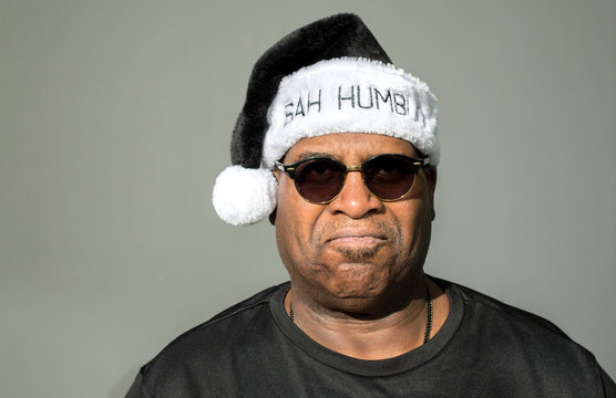 grumpy African American man wearing sunglasses and a black and white Bah Humbug hat isolated on a solid background