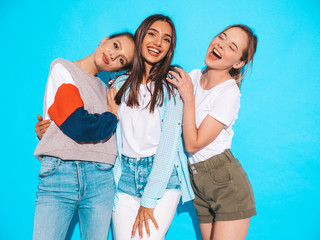 Three young beautiful smiling hipster girls in trendy summer clothes. Sexy carefree women posing near blue wall in studio. Positive models having fun and hugging