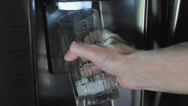 Man filling drinking glass with ice cubes from refrigerator ice cube dispenser. Stainless steel kitchen appliance with built in ice maker.