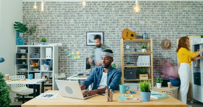 Time-lapse of African American man working with modern laptop in office while colleagues men and women are doing job in background. Creative people and work concep.