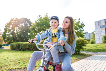 Mom woman with little boy son of 4-5 years old, learn to ride bicycle, have fun laughing and enjoy their vacation, summer autumn in city park site. Casual wear denim cap, sneaker pants.